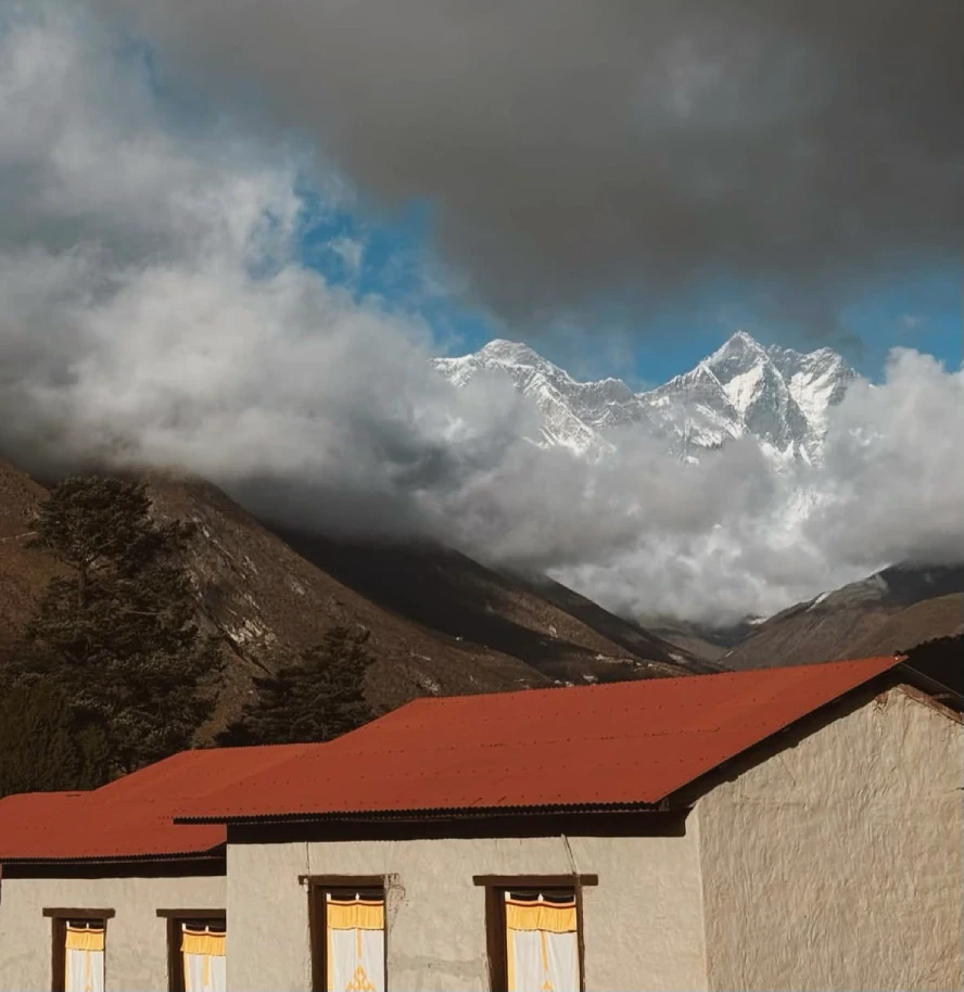 Mountain lodge with red roof beneath cloud-covered snow-capped Himalayan peaks in Nepal's Everest region.