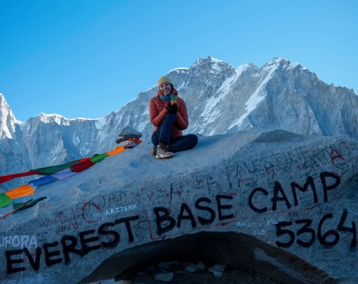 Smiling trekker at the Everest Base Camp sign with snow-capped Himalayan mountains and vibrant prayer flags along the Everest route in Nepal.