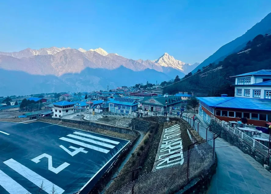 Lukla Airport runway overlooking the vibrant mountain town of Lukla, surrounded by traditional buildings and breathtaking snow-capped Himalayan peaks under a clear blue morning sky.