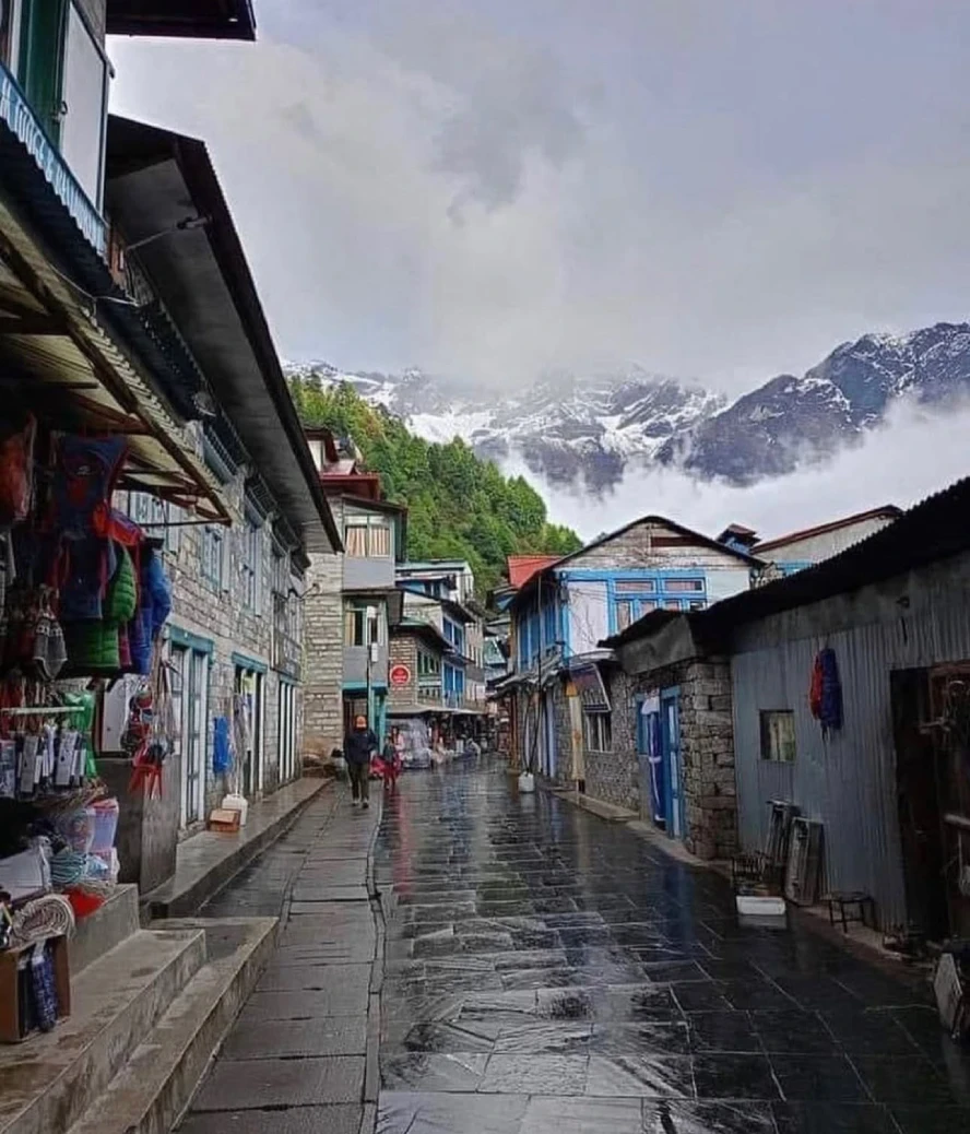 A rain-soaked Himalayan village street lined with traditional stone buildings and trekking shops, leading toward mist-covered, snow-capped mountains beneath a cloudy sky.