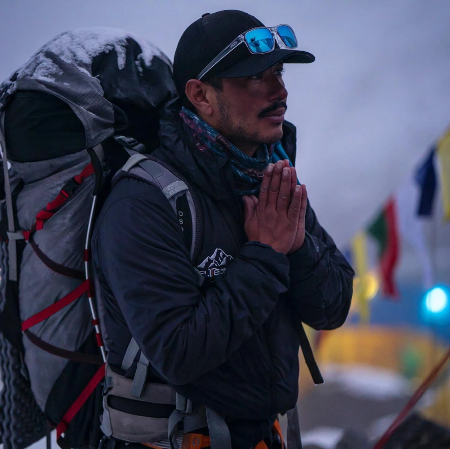 Nirmal Purja wearing mountaineering gear and carrying a large backpack during a Himalayan expedition, with his hands joined in a traditional Namaste gesture.