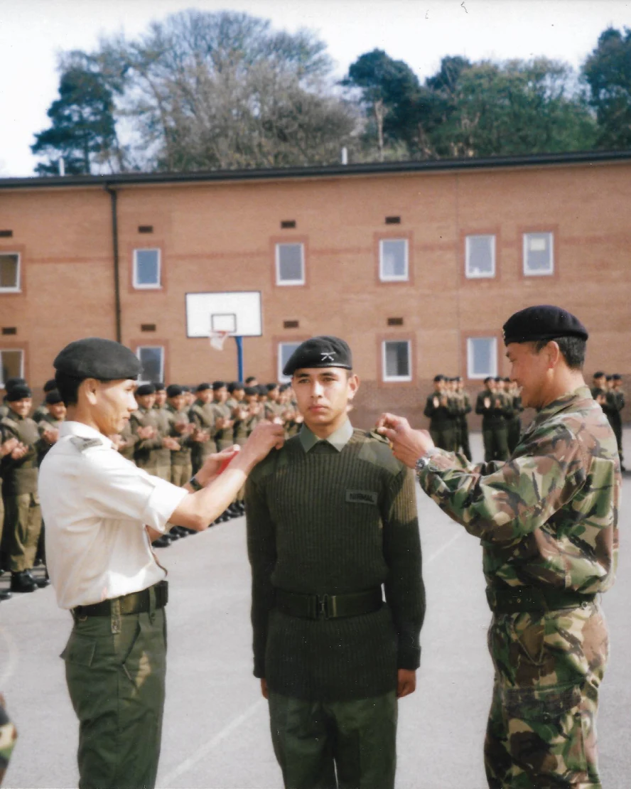 Archival photograph showing a young soldier standing during a military ceremony, with fellow soldiers gathered in formation in the background.