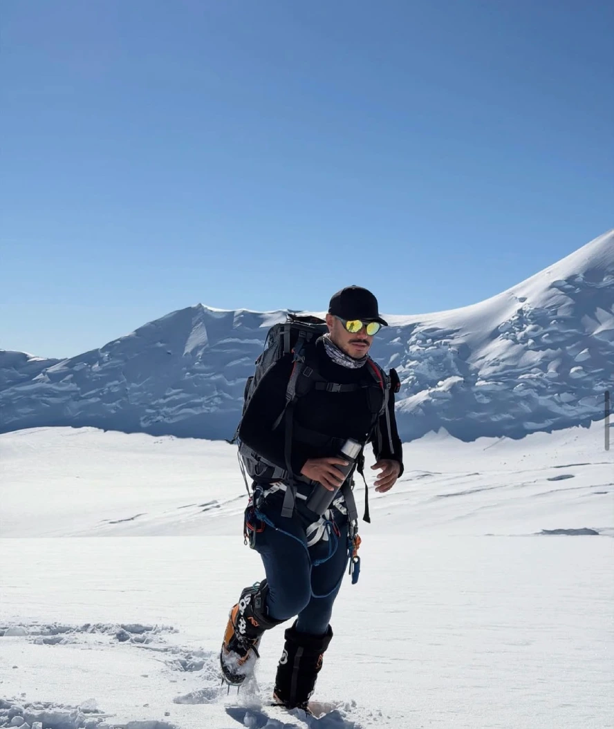 Nirmal Purja wearing high-altitude mountaineering gear and carrying a backpack while trekking across a snow-covered Himalayan landscape, with a glacier-covered mountain ridge in the background.