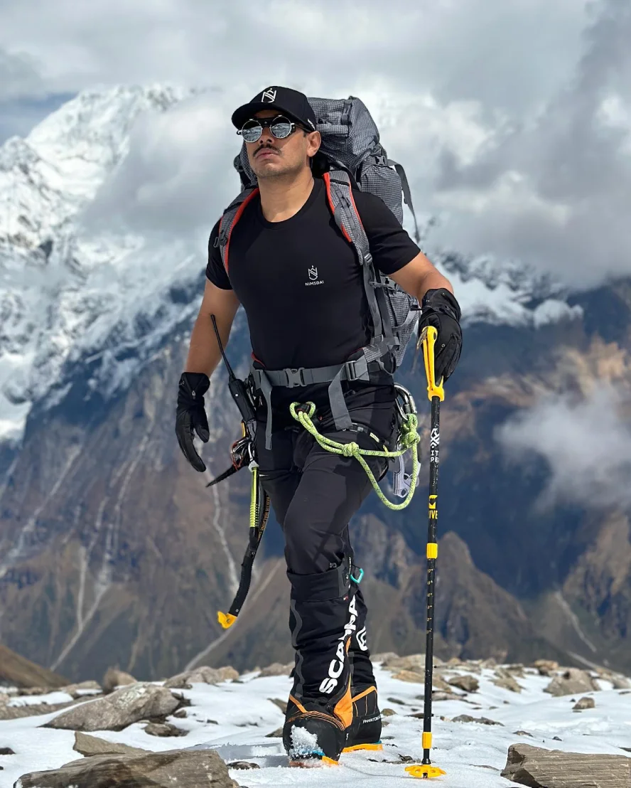 Nirmal Purja wearing mountaineering equipment, carrying a large backpack and trekking poles while crossing a snowy Himalayan landscape with dramatic mountain peaks in the background.