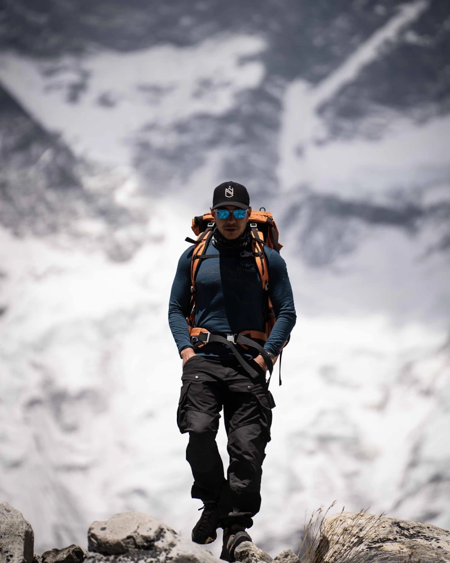 Nirmal Purja wearing mountaineering gear and carrying a backpack while trekking through a rocky Himalayan landscape with snow-covered mountains in the background.