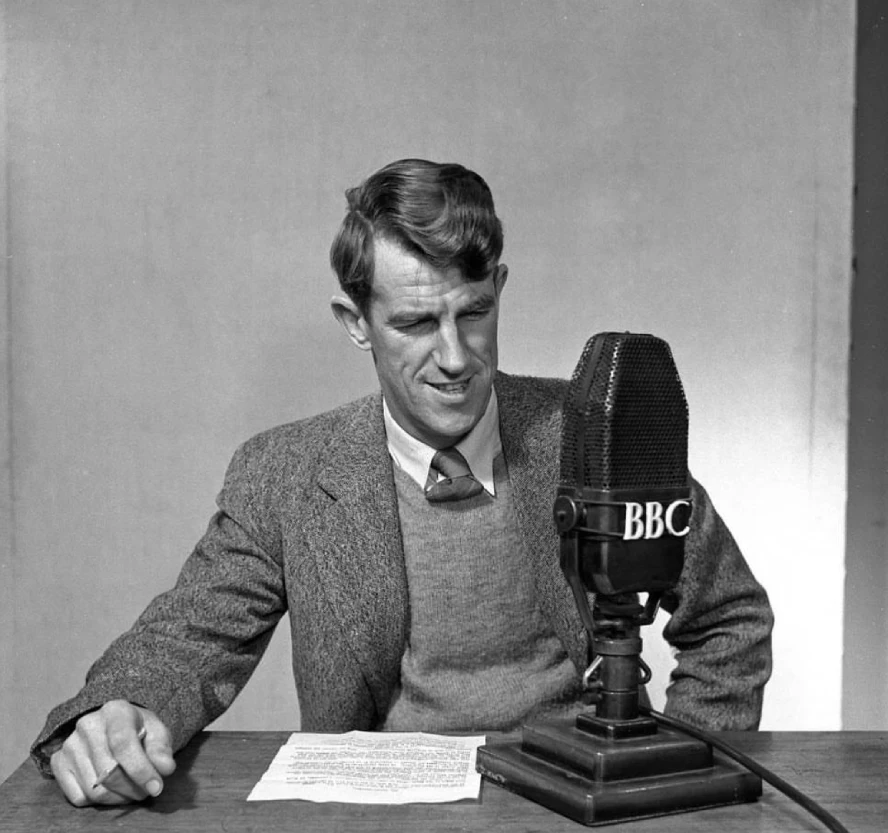 Black-and-white portrait of Sir Edmund Hillary speaking into a BBC microphone during a radio interview, dressed in formal attire shortly after his historic Mount Everest expedition.