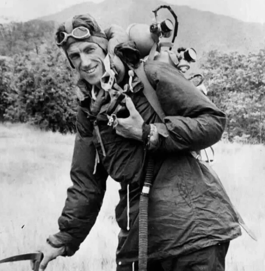 Black-and-white photograph of Sir Edmund Hillary wearing early mountaineering gear, carrying an oxygen set and climbing equipment during a Mount Everest expedition, with rugged hills in the background.