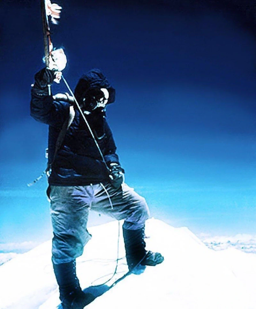 Historic image of Sir Edmund Hillary standing on the snow-covered summit of Mount Everest in full mountaineering gear, holding an ice axe with expedition flags against a deep blue Himalayan sky.