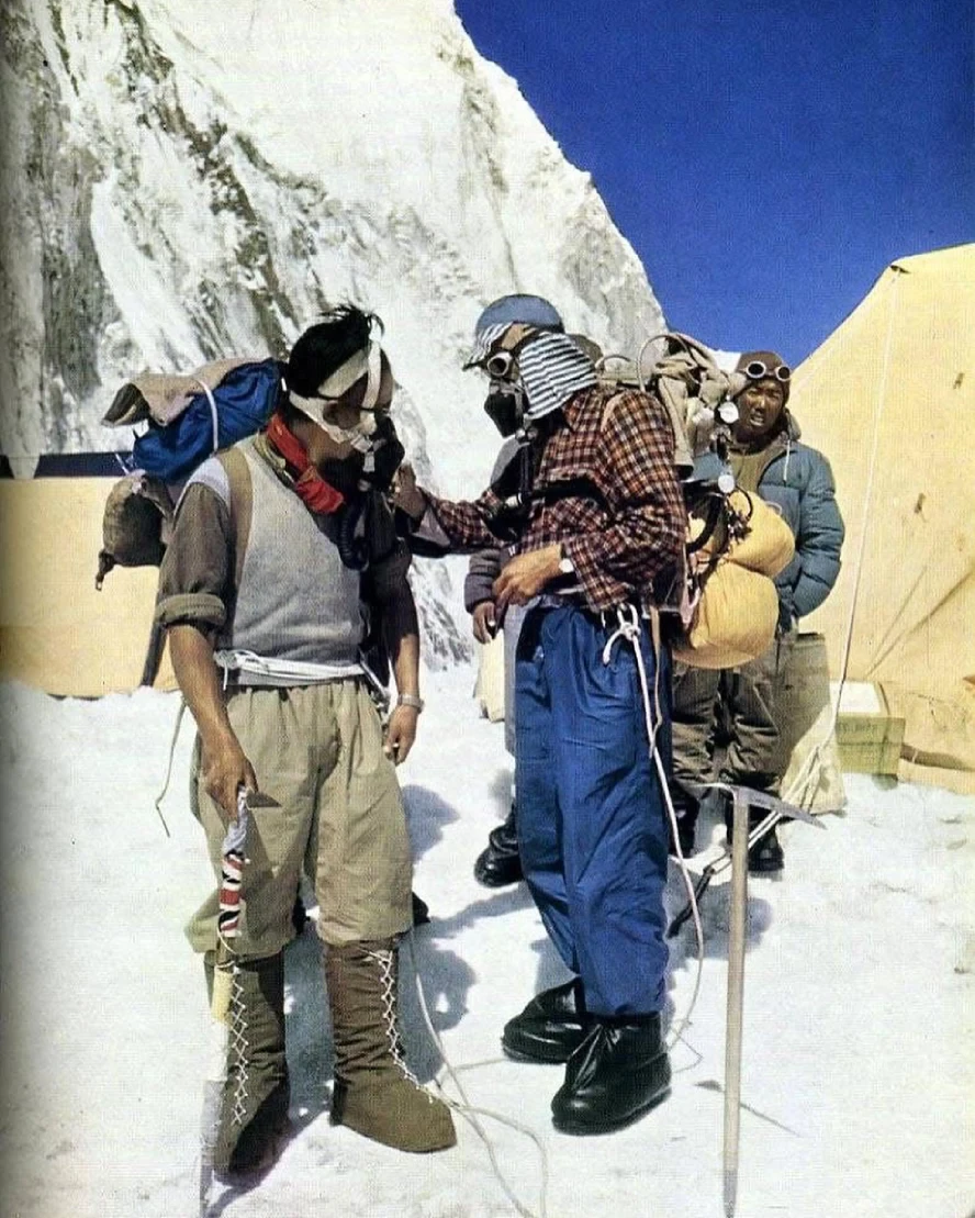 Tenzing Norgay Sherpa and fellow Everest climbers at a snowy high-altitude camp, wearing oxygen equipment and mountaineering gear beside expedition tents beneath towering Himalayan ice walls.
