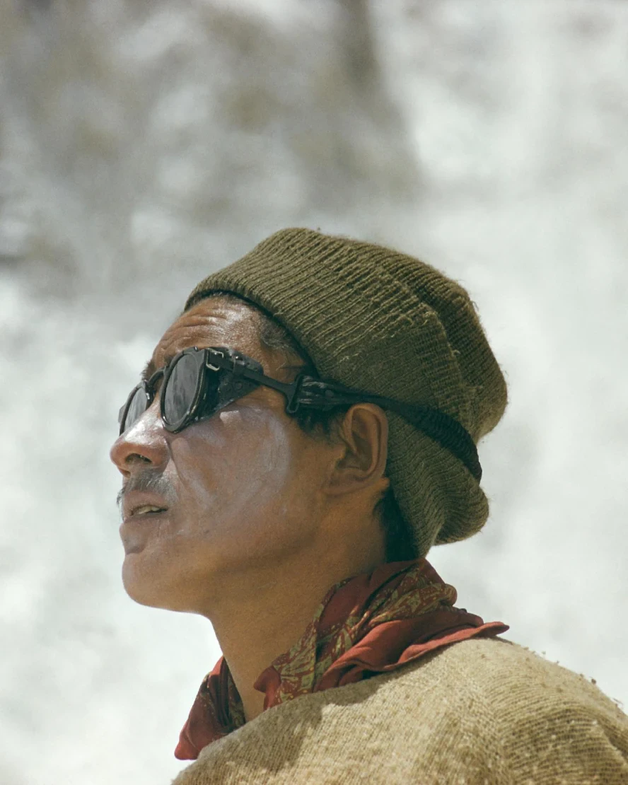 Portrait of Tenzing Norgay Sherpa wearing glacier sunglasses, a wool cap, and traditional expedition clothing, standing against a snowy Himalayan backdrop during a Mount Everest expedition.